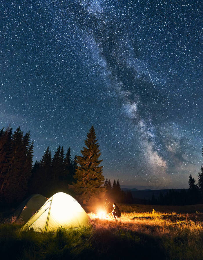 一个孤独的人坐在篝火边，在星空下的群山中。夜空中布满了明亮的星星和醒目的银河.山谷中的旅游帐篷，有高大的松树.