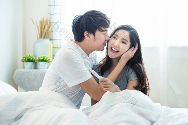 年轻的亚洲男子和妇女在家中卧床时，手里拿着一份快乐而快乐的妊娠期测试呈阳性.
