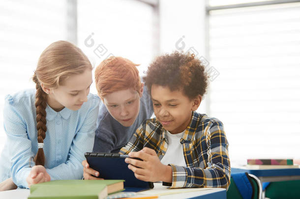 使用数字平板电脑或互联网的多族裔儿童坐在教室复制空间的课桌前