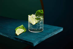 带有新鲜饮料的老式玻璃杯，薄荷叶和石灰片，蓝色木制表面，绿色和蓝色几何背景