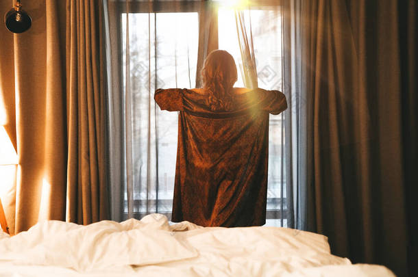 一个女人在卧室的窗前遇见了黎明，她穿着丝绸长袍拉开窗帘