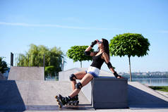 年轻漂亮的黑发女子，身穿旱冰鞋和安全设备，坐在城市湖边的金属滑翔机平台上。夏季滑冰鞋运动健美姑娘的全长坐姿画像