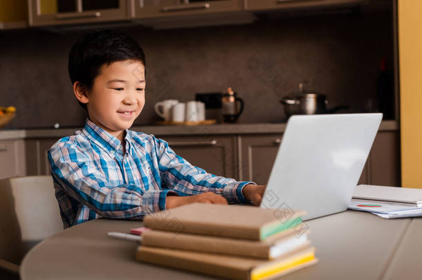 在隔离期间，带着笔记本电脑和书籍在<strong>网上学习</strong>的亚洲男孩微笑着