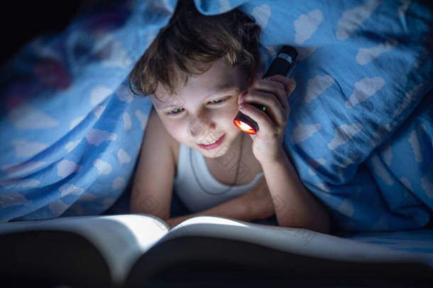 快乐的男孩躺在毯子下，用手电筒在黑暗中看书，而不是在每个人都睡着的时候睡觉。那孩子躲得远远的对知识的渴望.