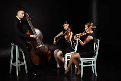 三位专业音乐家在小提琴上演奏古典音乐，在黑暗舞台上演奏对比度低音