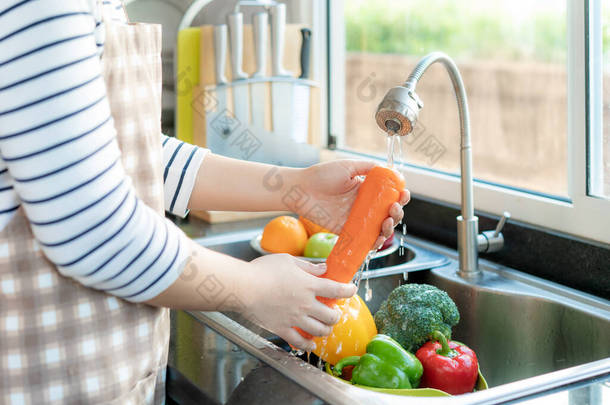 亚洲健康妇女在<strong>厨房水池</strong>上方清洗胡萝卜和其他蔬菜，用水清洗水果/蔬菜，以消除污染机会