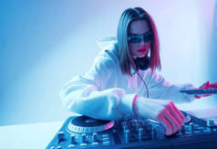 年轻酷女孩DJ在混合控制台和笔记本电脑上混合着音乐，穿着时髦的衣服，戴着霓虹灯背景的眼镜.