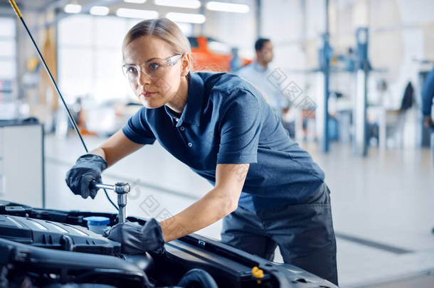 美丽的增强女性力量的机修工是在汽车服务公司的汽车上工作。戴安全眼镜的女人正在修理发动机.她用的是斧头。现代汽车清洁工场.