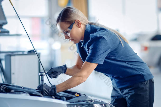 美丽的增强女性力量的机修工是在汽车服务公司的汽车上工作。戴安全眼镜的女人正在修理发动机.她用的是斧头。现代车辆洁净工场.