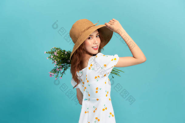 身穿<strong>夏装</strong>，头戴草帽的年轻美丽的亚洲女人，孤零零地站在蓝色的背景上，摆出一副花束的架势