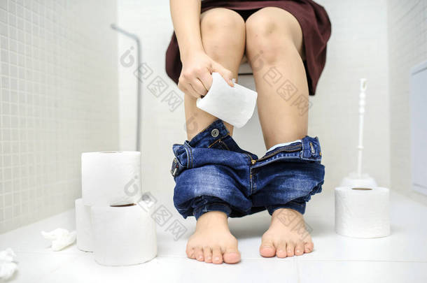 有便秘或腹泻的近照妇女<strong>坐在</strong>马桶上，裤腿下垂，<strong>地上</strong>有很多卫生纸卷