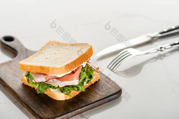 白大理石表面刀叉附近木制切菜板上带有牛油果和牛油果的新鲜三明治的精选焦点