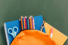 彩色铅笔、笔记本、剪子和画笔在绿色黑板附近的黄色背包的顶部视图
