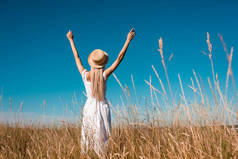 穿着白衣、头戴草帽的时髦女人站在青草丛生的草地上，高举双手在蓝天衬托下的背影