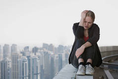 可悲的年轻女商人坐在摩天大楼的屋顶边缘，背后是城市的景象。工作压力和失败.