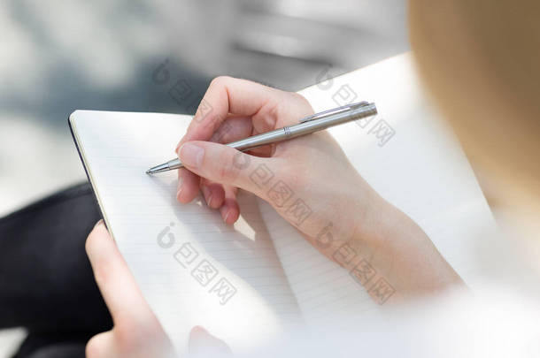 <strong>女性</strong>坐在室外用笔在空白日记本上用手写单子的服装.