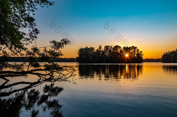 夏末在宾夕法尼亚的Pymatuning湖上落日。前景是一棵倒下的树，太阳升起来穿过背景岛上的树木，天空是橙色和蓝色的.