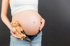 在怀孕妇女的身边，穿着拉链牛仔裤，抱着泰迪熊靠近她的腹部，背景五彩斑斓，有复制空间。期待一个婴儿的概念.