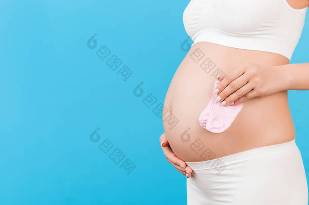 为孕妇手中的女婴剪下粉色袜子的图像，在蓝色背景下靠在她的腹部。父母身份的概念。复制空间.