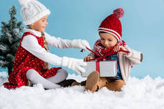 戴着帽子和冬装的孩子们坐在雪地上，触摸着被蓝色隔离的礼物