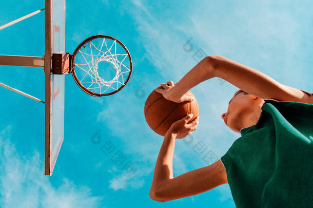 体育和篮球。一个身穿绿色运动服的少年把球扔进了篮筐。底部的观点。背景是蓝天.