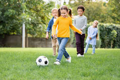 在公园里与多民族朋友一起踢足球的女孩在模糊的背景下嬉笑