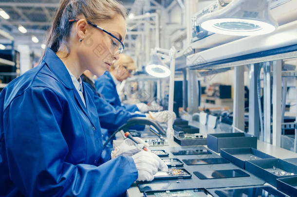 蓝色工作服和防护眼镜中的女性电子工厂工人正在用螺丝刀组装智能手机。拥有更多员工背景的高科技工厂设施.