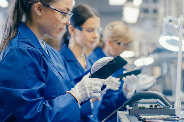 蓝色工作服和防护眼镜中的女性电子工厂工人采用智能手机屏风并进行质量检查。拥有多名员工的高科技工厂设施.