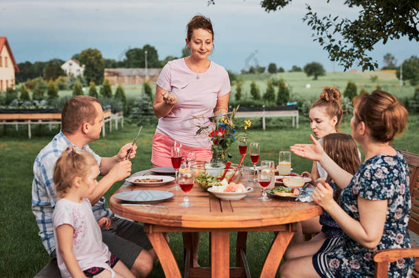 夏天外出野餐时,<strong>一家人</strong>在自家花园的烤架边<strong>吃饭</strong>.围坐在餐桌旁，端着食物和碗碟的人