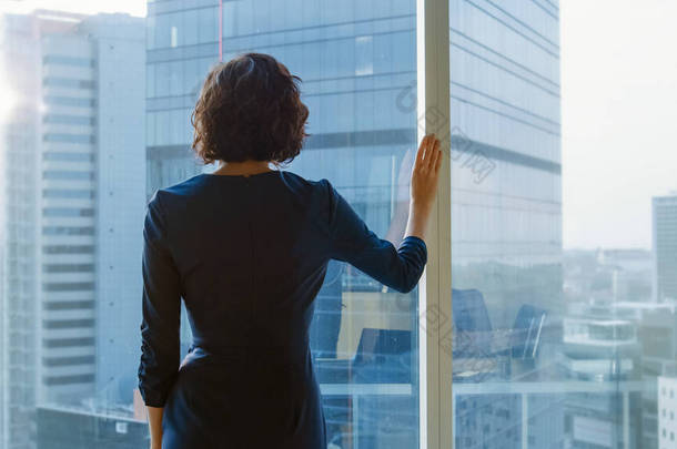拍的成功的女商人在一个罢工的黑色衣服在她的办公室里凝视着窗外沉思。具有个人电脑和大城市视野的现代商务办公室.