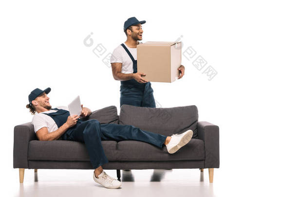 快乐的劳动者躺在沙发上，带着数码平板电脑，接近快乐的印第安人搬运工，手里拿着白色的盒子