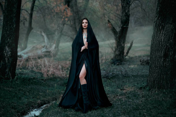 神秘的幻想哥特式女人被邪恶缠住了。穿着黑衣斗篷的女恶魔吸血鬼。走在<strong>密密麻麻</strong>的森林深处，树木丛生。中古女王穿著丝绸斗篷，围巾摆姿势