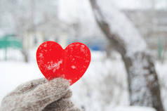 在城市公园、雪地、冬季、情人节的概念、特写等模糊的背景下，一个女人手戴着针织的温暖手套，手握一颗红心