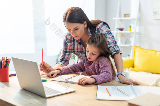 母亲和女儿在家里的书桌边看着笔记本电脑，背景模糊不清
