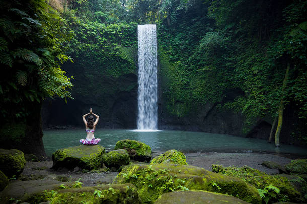 瑜伽莲花姿势。年轻的白人妇女坐在石头上，沉思着练习瑜伽，在瀑布边弹跳。手举起来在namaste mudra 。瑜伽<strong>撤退</strong>。从后面看蒂布马纳瀑布巴厘