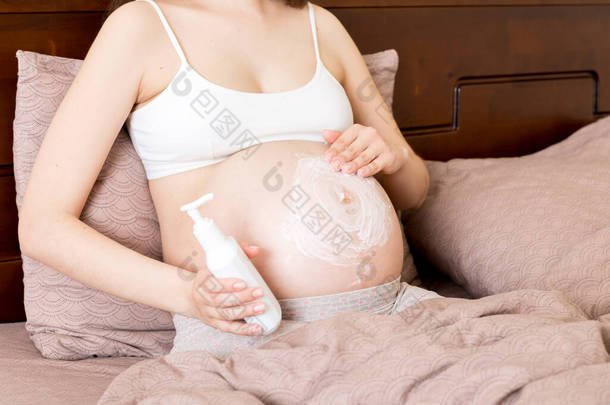 一个怀孕的女孩坐在家里的床上，涂片者笑着在她的肚子上涂了抗<strong>拉伸</strong>标记霜。怀孕、生育、准备和期望概念.