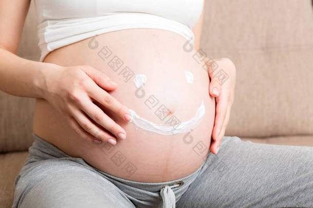 一个怀孕的女孩<strong>坐在</strong>家里的<strong>床上</strong>，涂片者笑着在她的肚子<strong>上</strong>涂了抗拉伸标记霜。怀孕、生育、准备和期望概念.