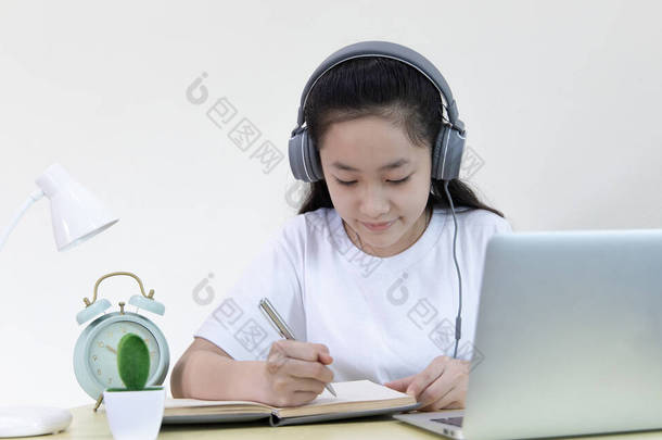 亚洲女孩在笔记本电脑上学习，并在笔记本上写着打算、在线学习、待在家里、新常态、 Covid-19 Coronavirus 、社交疏远、网上学习