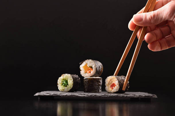 背景手在<strong>黑色</strong>背景的深色木桌上，用筷子在板子上采摘4个不同种类的蔬菜寿司。前面的景色1.横向构成.