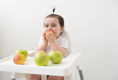 坐在婴儿椅上的女婴吃苹果，在白色背景下微笑。婴儿第一固体食物