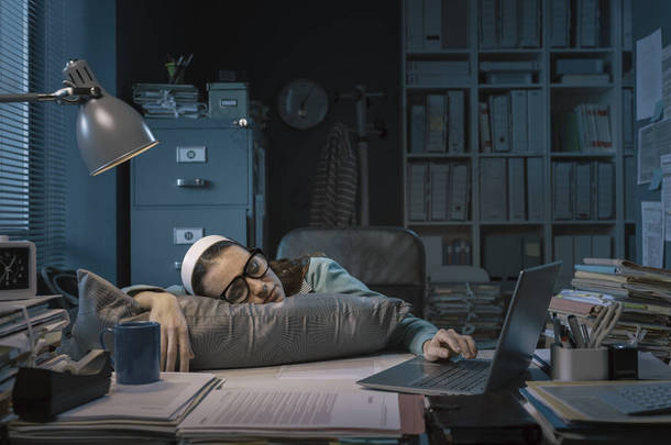 Exhausted young office worker sleeping at her desk, job burnout and overtime work concept