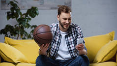 快乐的篮球迷拿着球，用智能手机观看比赛