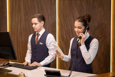 两个年轻的酒店接待员中的一个站在柜台旁，看着触摸板展览，并与同事在电话上咨询客户意见