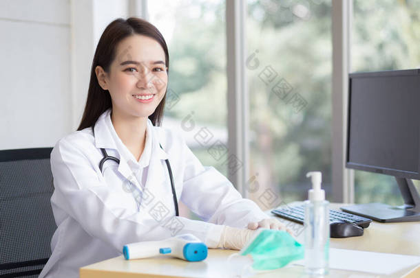 亚洲漂亮的年轻微笑的女<strong>医生戴</strong>着橡胶<strong>手套</strong>坐在医院的办公室里。桌上有一张纸、面罩、酒精凝胶、温度计和电脑.