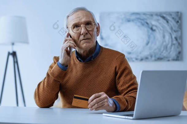 一个严肃的老人，坐在笔记本电脑旁拿着信用卡，在家里用手机聊天