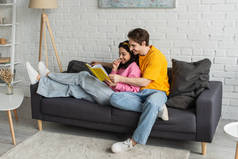 幸福的年轻夫妇躺在沙发上，抱着书，在客厅里看书