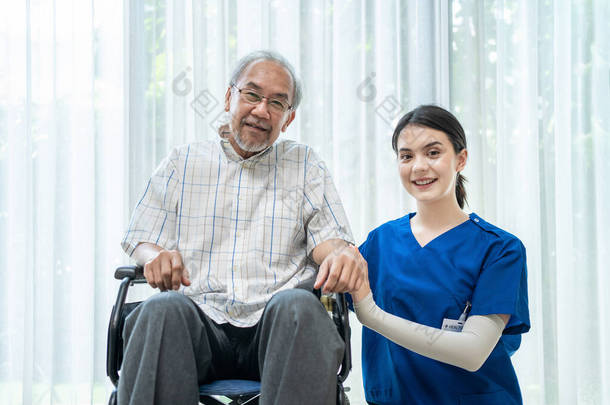 坐在轮椅上的快乐的亚洲残疾老年男性的肖像享受呆在家里的护理。漂亮的<strong>看护</strong>护士的女人坐在老年<strong>病人</strong>旁边，看着镜头。医疗保险概念.