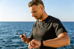 英俊的成年男子使用智能手机和手表在海滨