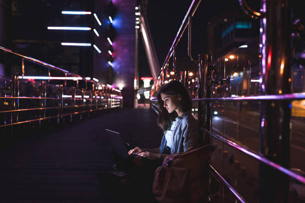 年轻女子的侧面视图使用膝上型电脑在街道上与夜间城市灯光背景