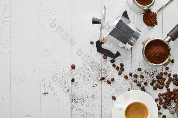 在白色木质表面与咖啡豆一起享用的门廊、勺子和咖啡杯附近的间歇泉咖啡机的顶视图